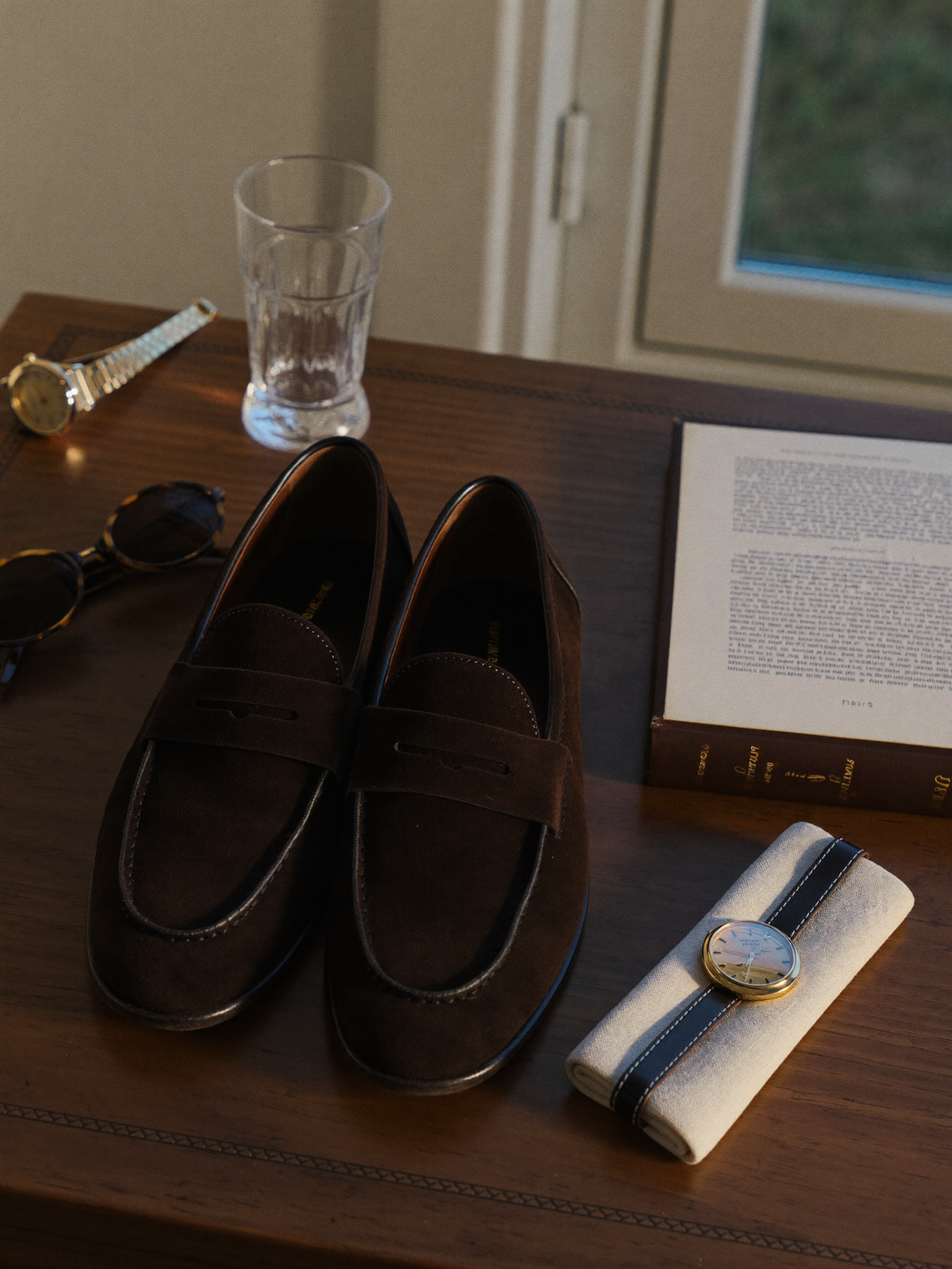 Suede loafers, watch and sunglasses on a walnut desk